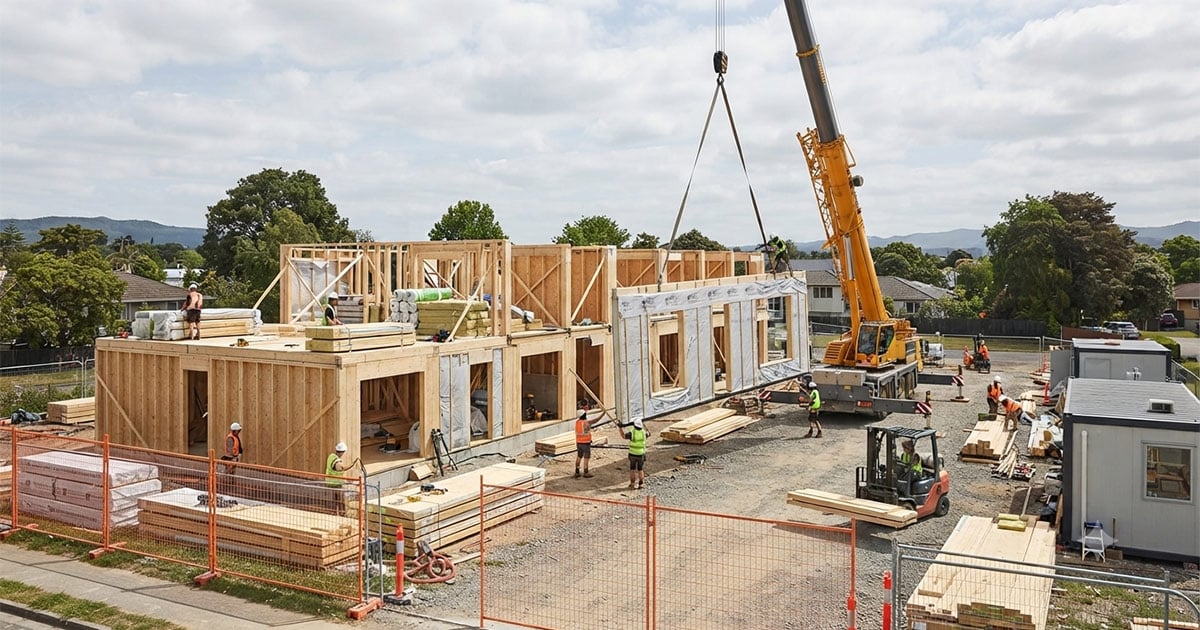 Crane lifting a prefabricated timber-frame wall panel into place during house assembly on a construction site.
