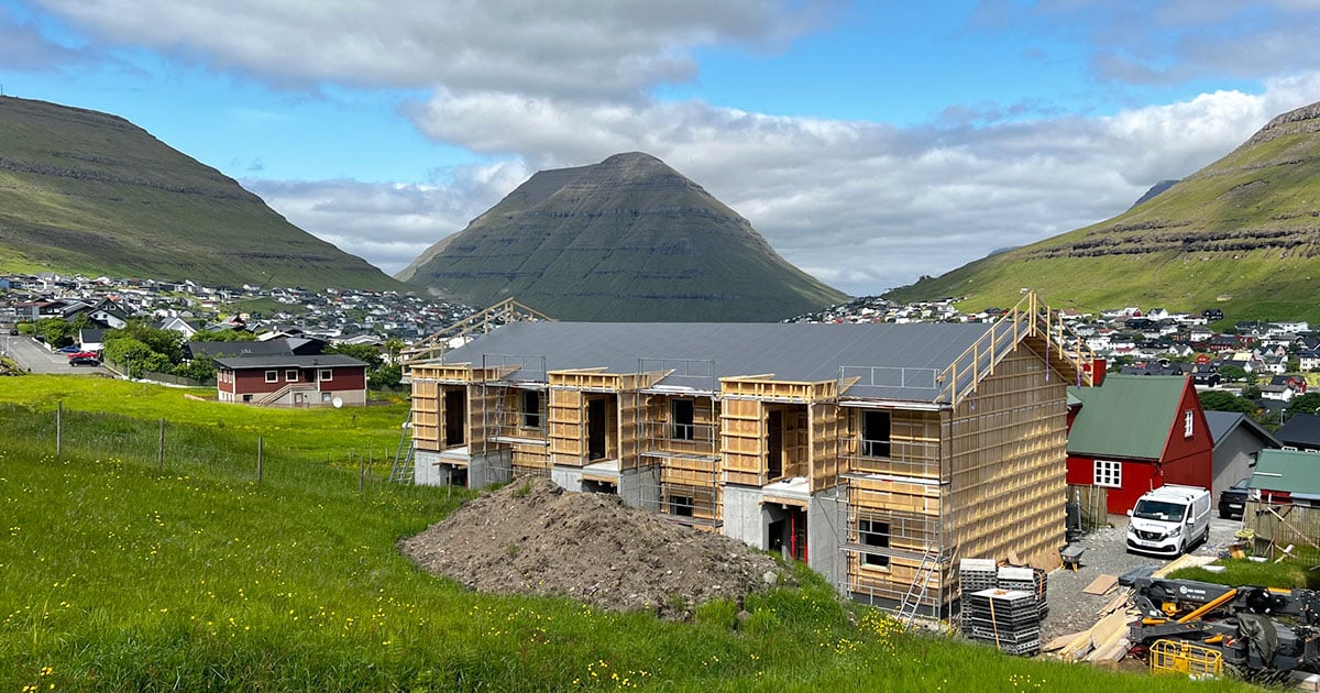 Three-apartment timber-frame building under construction in the Faroe Islands with mountains and village in the background.