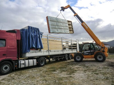 Prefabricated timber-frame wall panel being lifted from a truck by a crane on site.